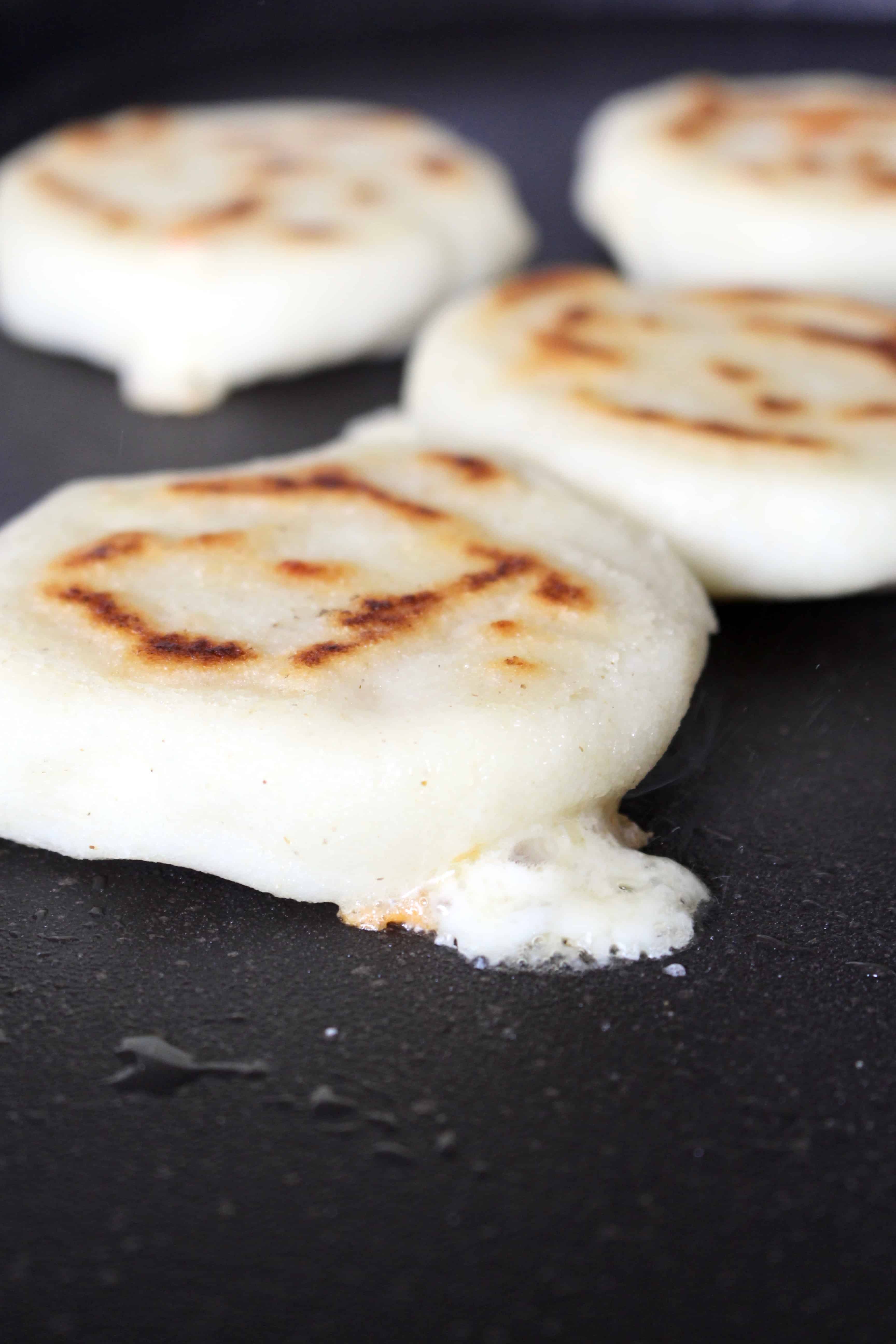 Cheese oozing out of an arepa cooking on a black griddle.