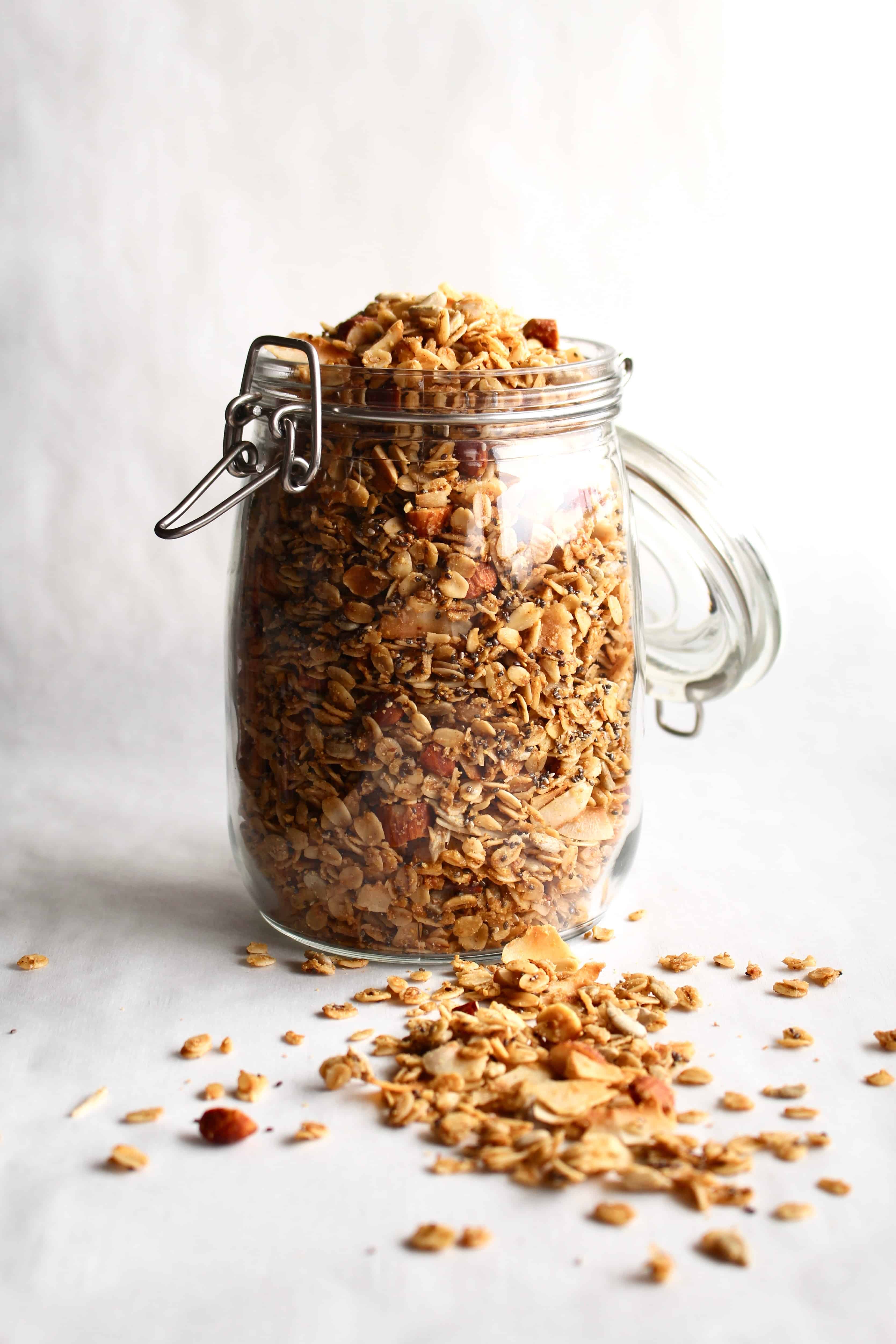A glass jar of gluten-free granola on a white surface, with some granola spilled in front of the jar.