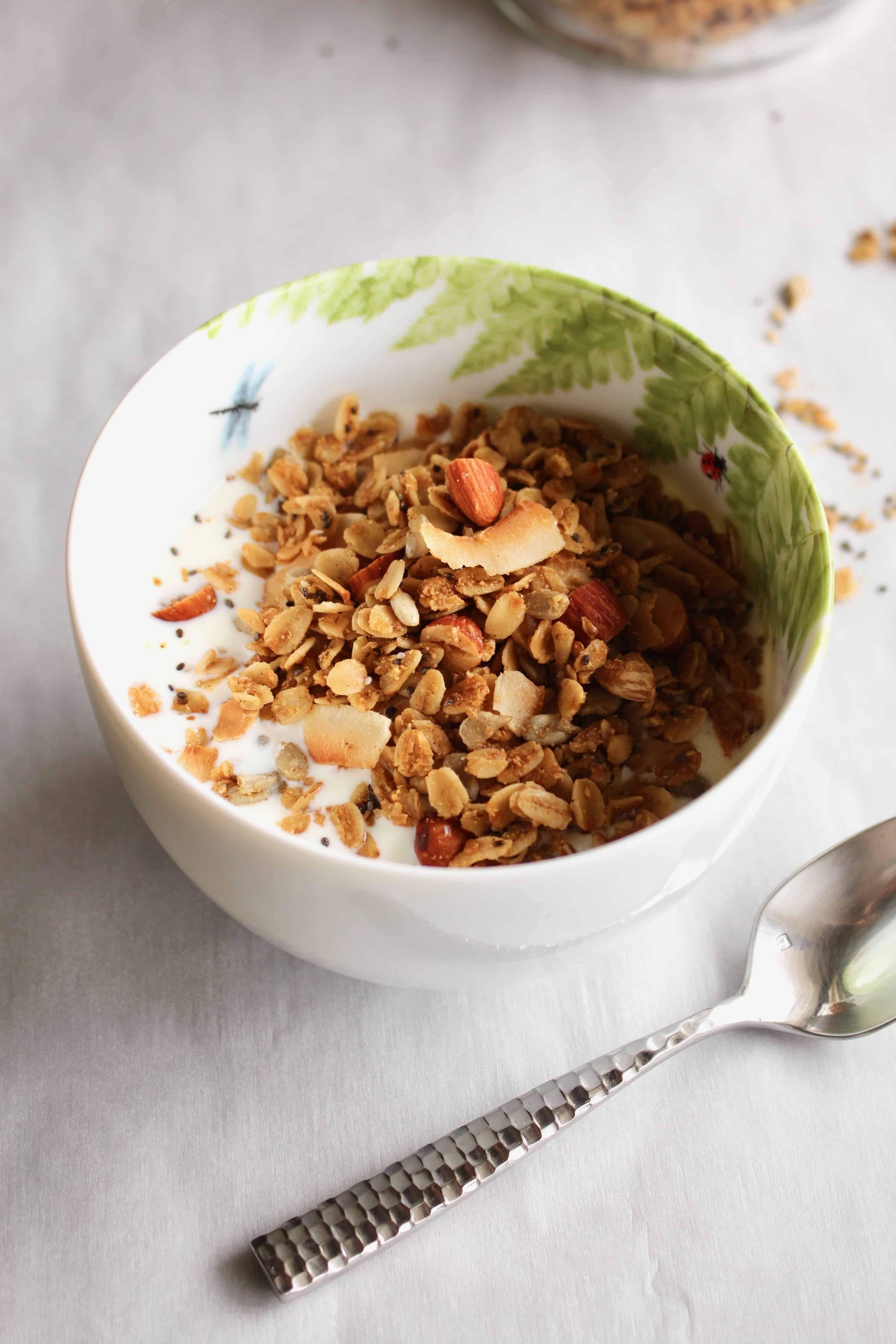 A white bowl with gluten free granola and milk, with a silver spoon next to it.