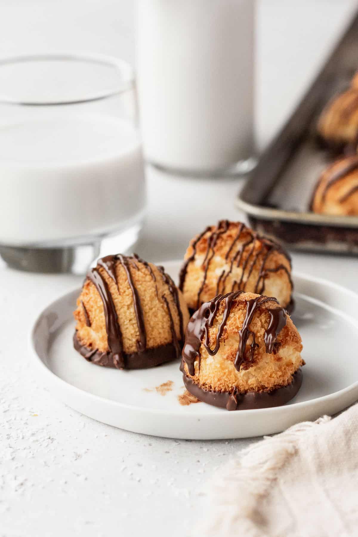 3 chocolate drizzled coconut macaroons on a white plate with a glass of milk in the background.