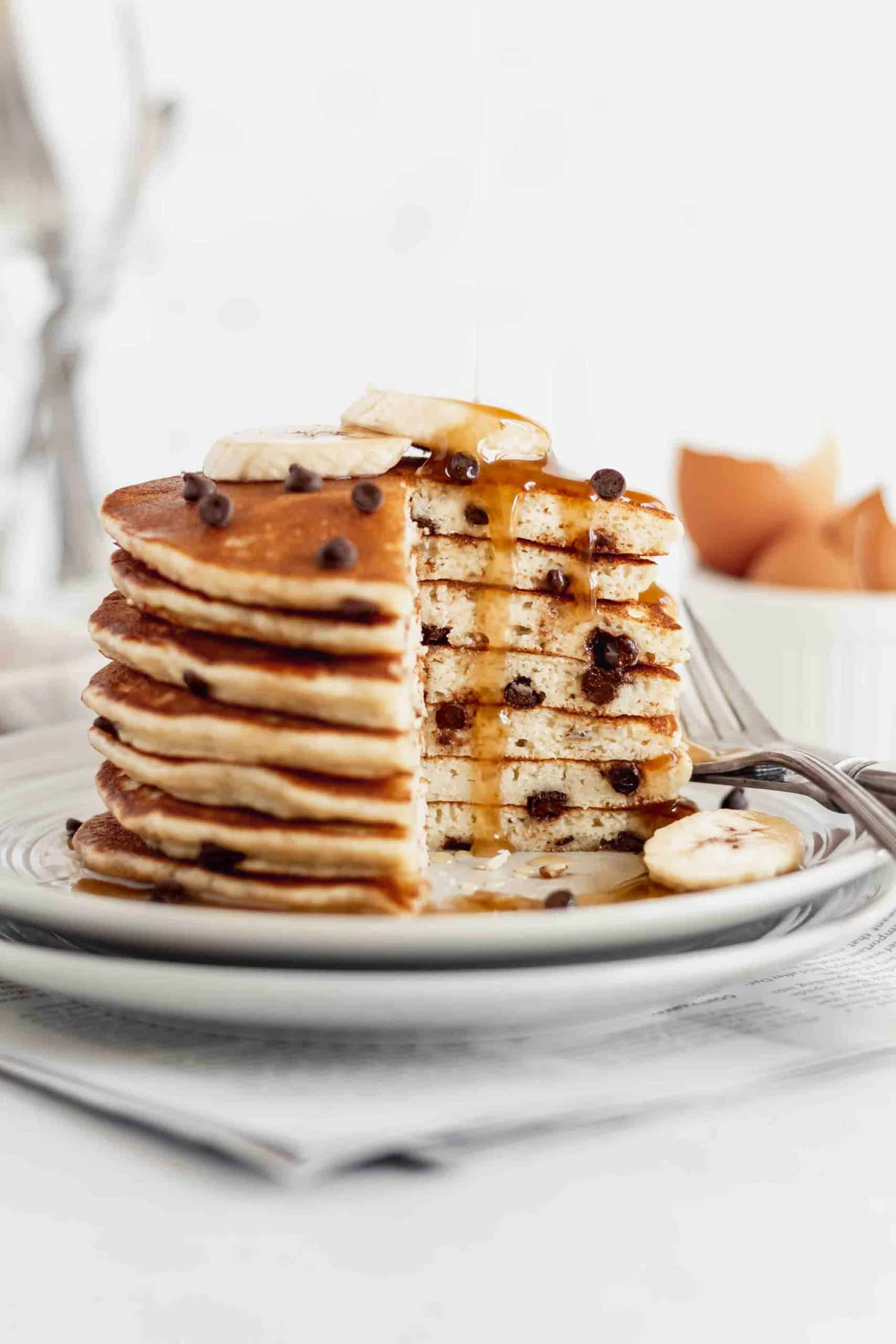 A stack of gluten free banana pancakes with chocolate chips, with maple syrup getting drizzled on.