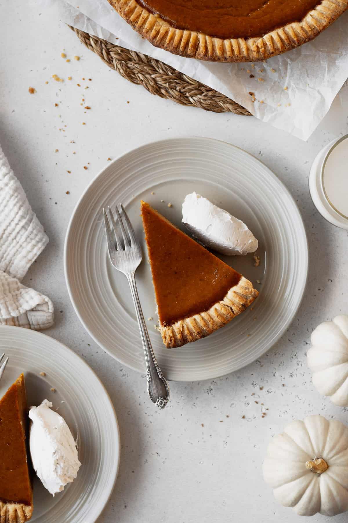 Overhead shot of a slice of dairy-free pumpkin pie with whipped cream and a fork on a plate surrounded by plates and white pumpkins.