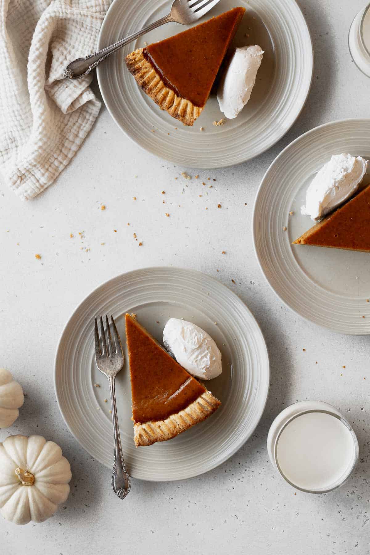 Overhead shot of 3 dairy-free pumpkin pie slices on plates with whipped cream and a couple forks.