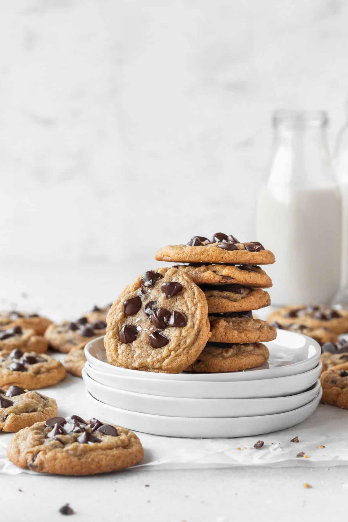 stack of gluten free chocolate chip cookies on a stack of white plates with a glass jug of dairy-free milk in the background.