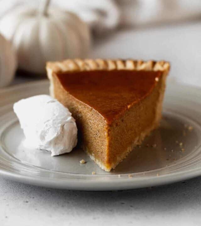 A slice of dairy-free pumpkin pie on a grey plate with a whipped coconut cream on the side and a white pumpkin in the background.