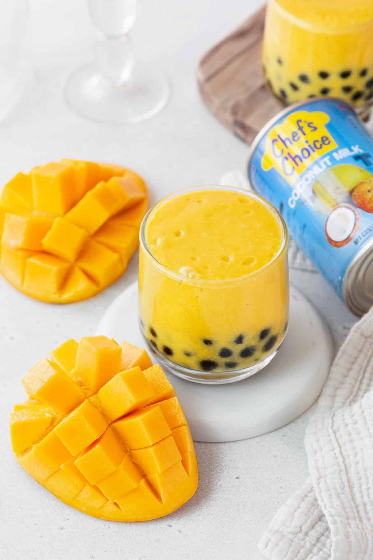 A small glass of the mango boba tea next to a can of chef's choice coconut milk and a cubed mango.