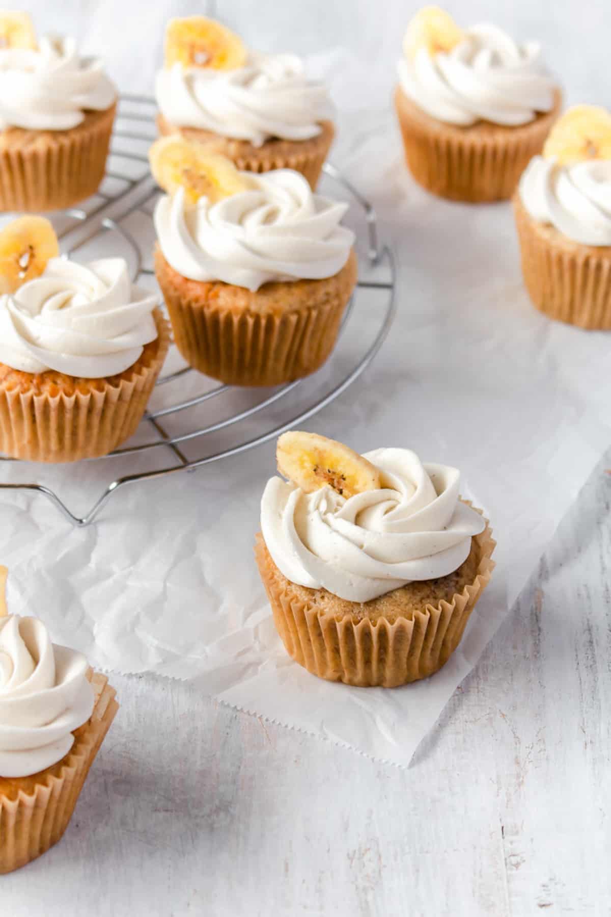 Banana cupcakes with buttercream frosting piped on top and decorated with a banana chip scattered on a white surface.