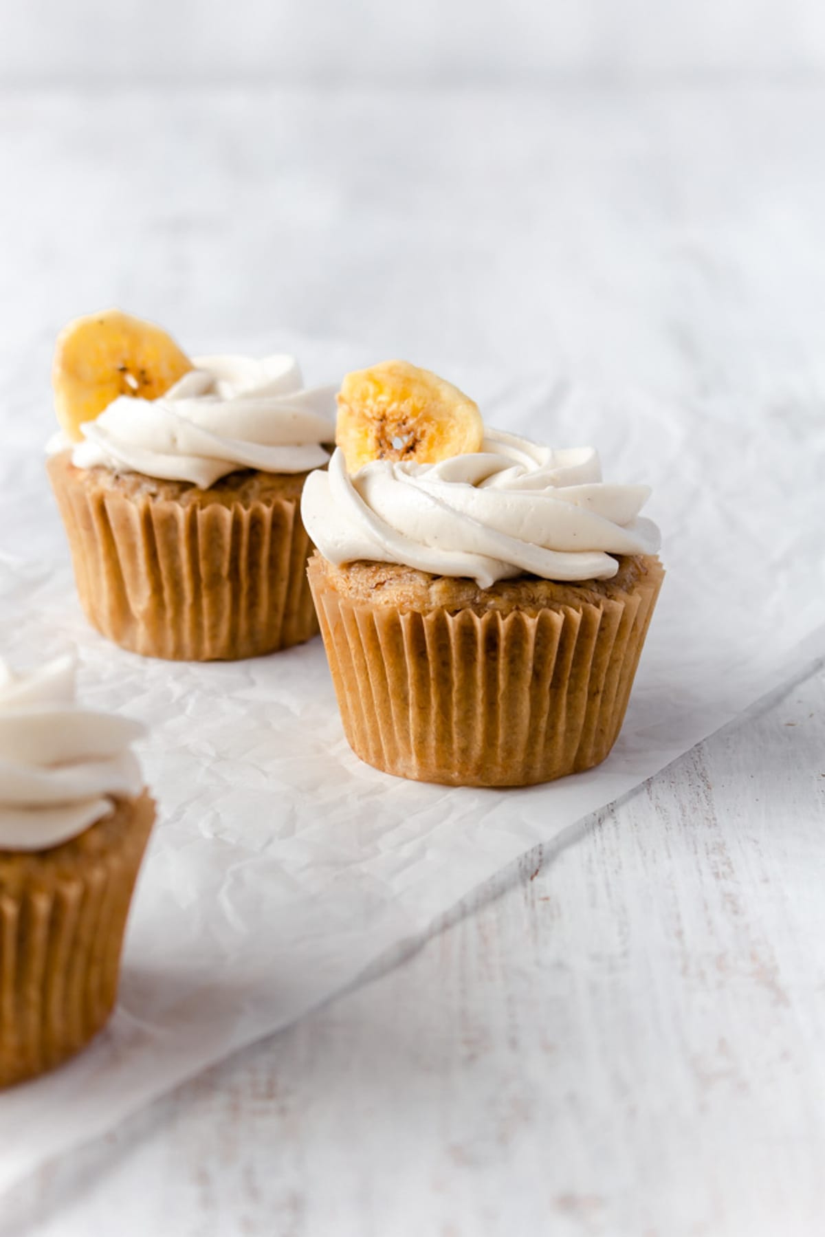 Two banana cupcakes on a piece of parchment paper with another cupcake off to the side.