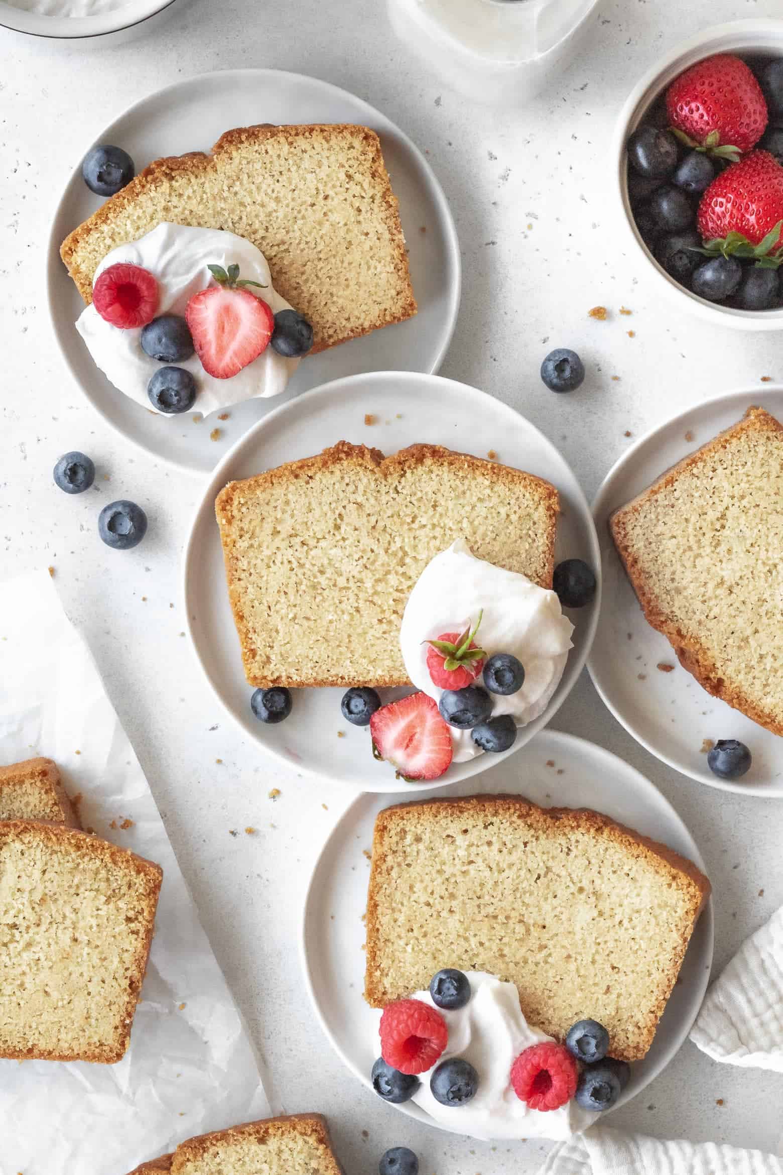 Slices of gluten free pound cake on small white plates with coconut cream and berries.