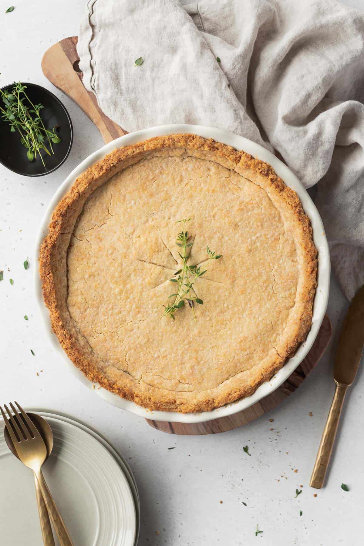 A baked plant based chicken pot pie with sprigs of thyme on top with gold utensils and a linen around it.