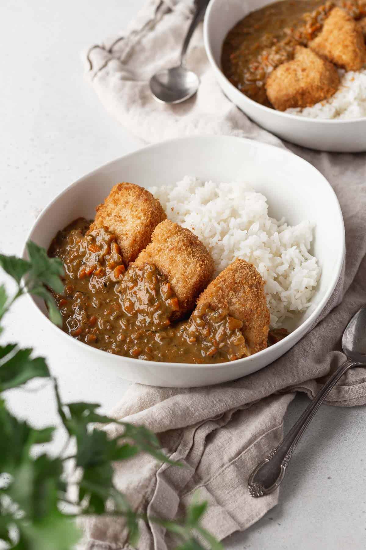 Vegan katsu curry in a white bowl on a linen with a flat leaf parsley plant in the corner of the shot.