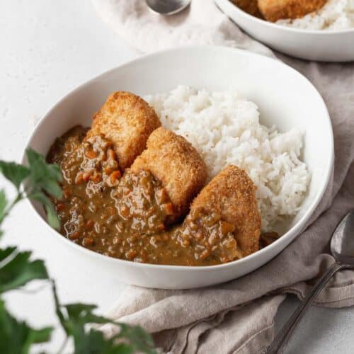 Vegan katsu curry in a white bowl on a linen with a flat leaf parsley plant in the corner of the shot.