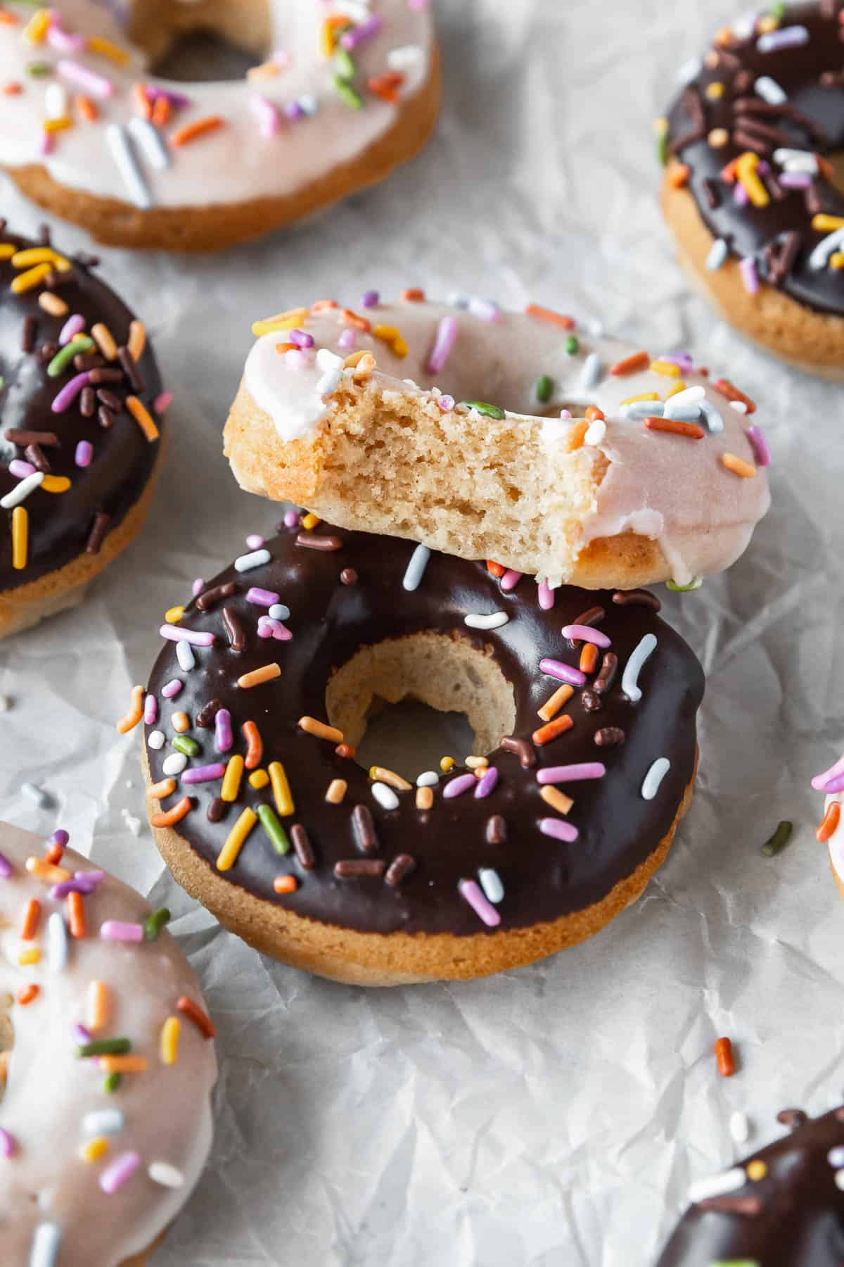 45 degree angle shot of a chocolate glazed and sprinkled donut with a vanilla glazed vegan doughnut with a bite taken out to show the tender crumbed interior.