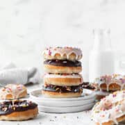 Stack of alternating chocolate and vanilla glazed baked vegan donuts on a stack of white dessert plates with a jug of non-dairy milk in the background.
