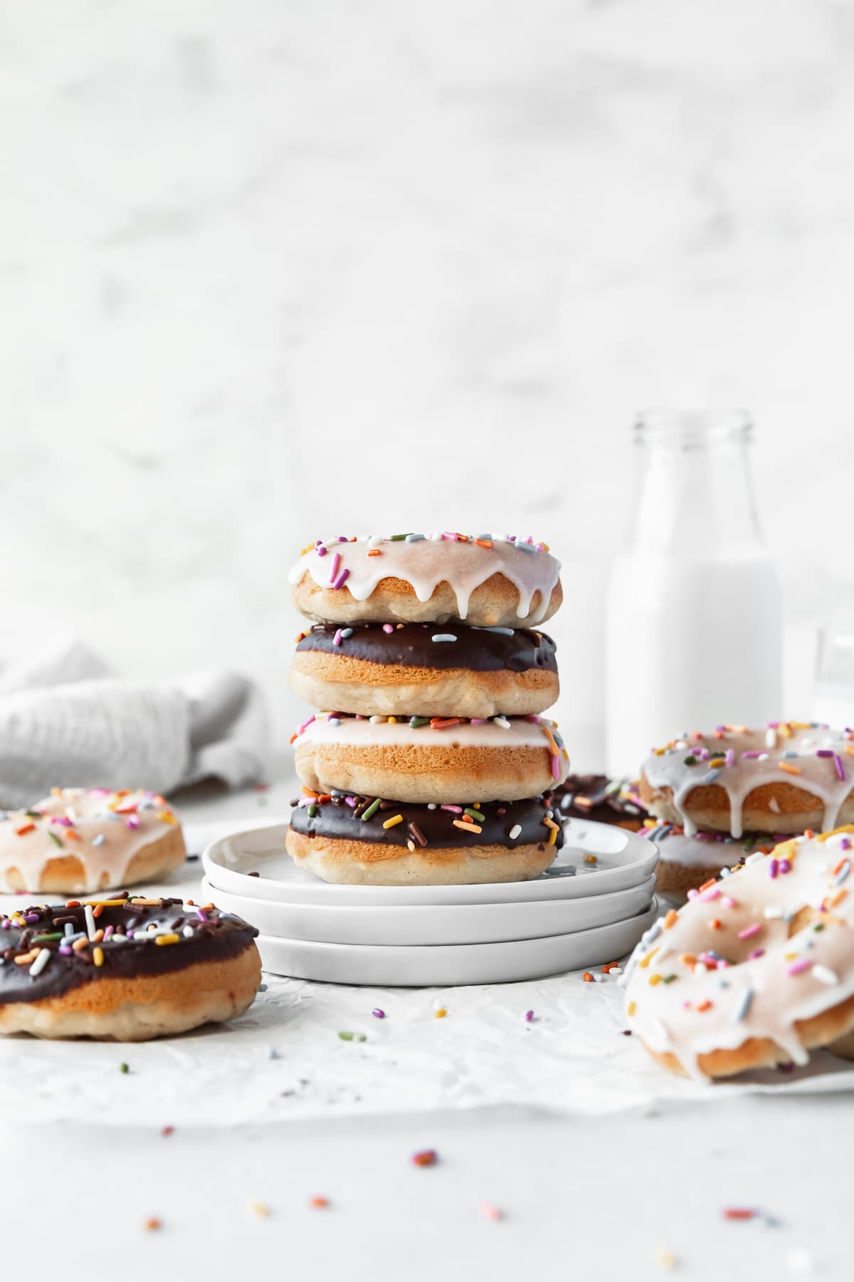 Stack of alternating chocolate and vanilla glazed baked vegan donuts on a stack of white dessert plates with a jug of non-dairy milk in the background.