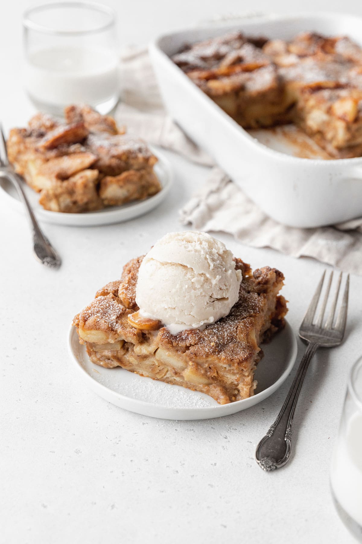 A slice of vegan bread pudding on a white plate, topped with vegan vanilla ice cream, with another plate of bread pudding and a white casserole dish in the back of the scene.