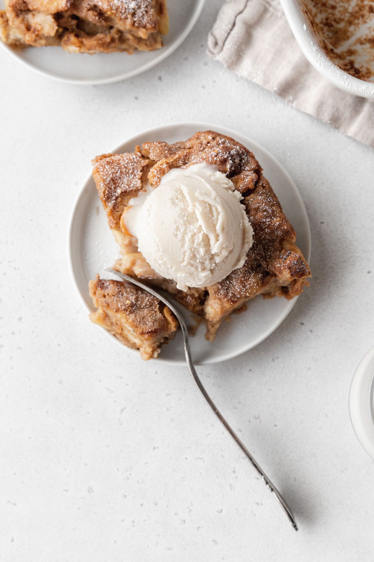 Overhead shot of a slice of vegan bread pudding topped with vanilla ice cream, with a silver fork cutting a bite off the corner.