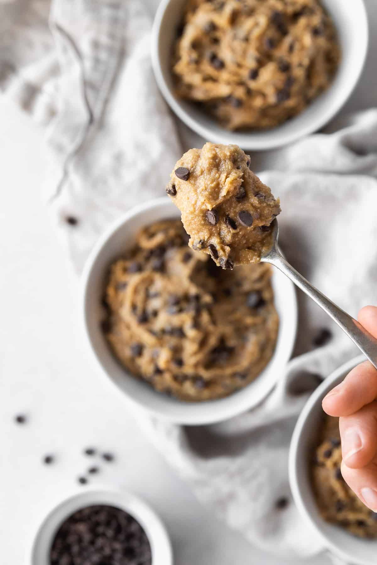 Closeup on a spoonful of edible vegan cookie dough with the three bowls filled with it blurred out in the background.