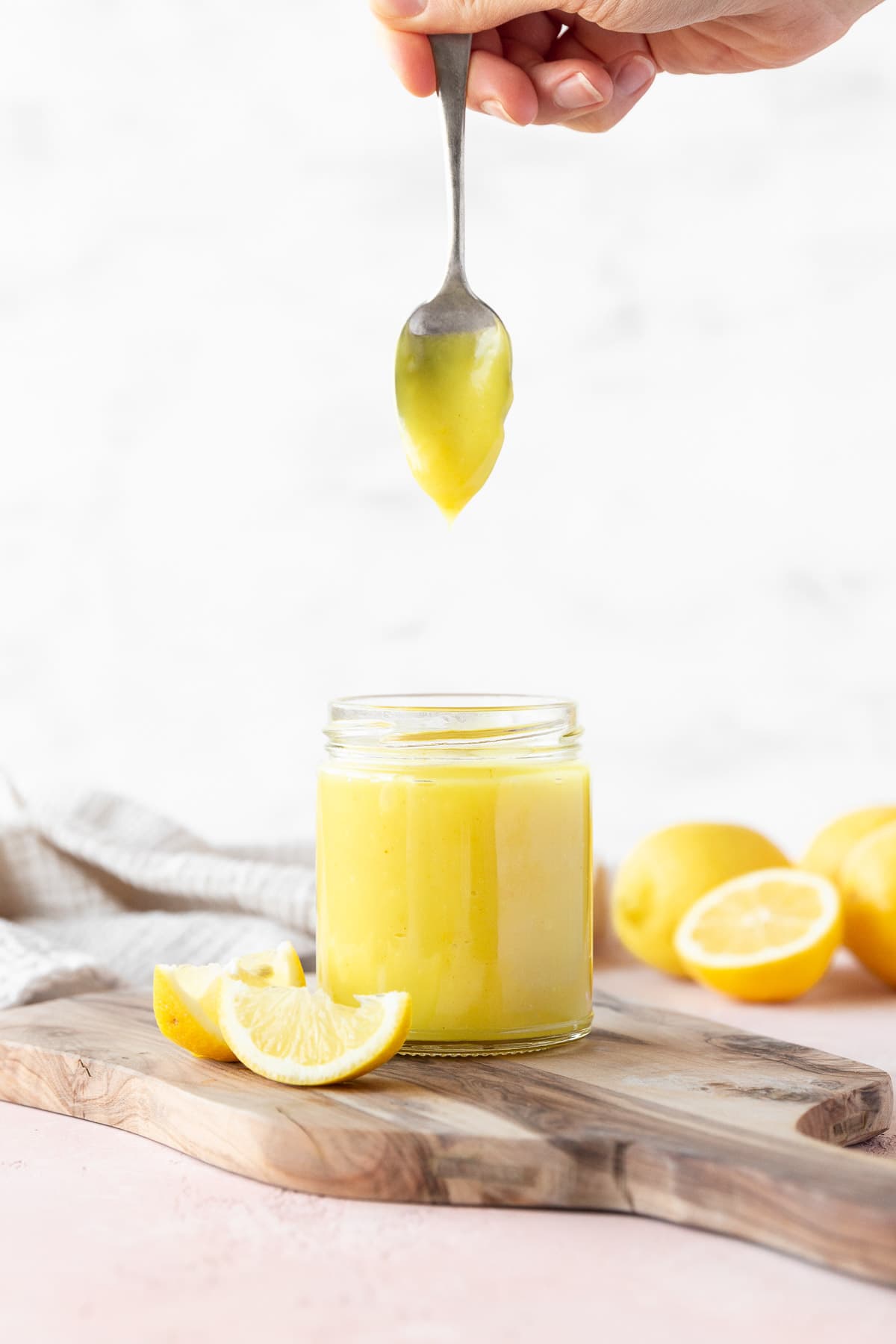 Side on shot of a hand holding a spoon of vegan lemon curd vertically over the full mason jar with the curd slowly dripping off.