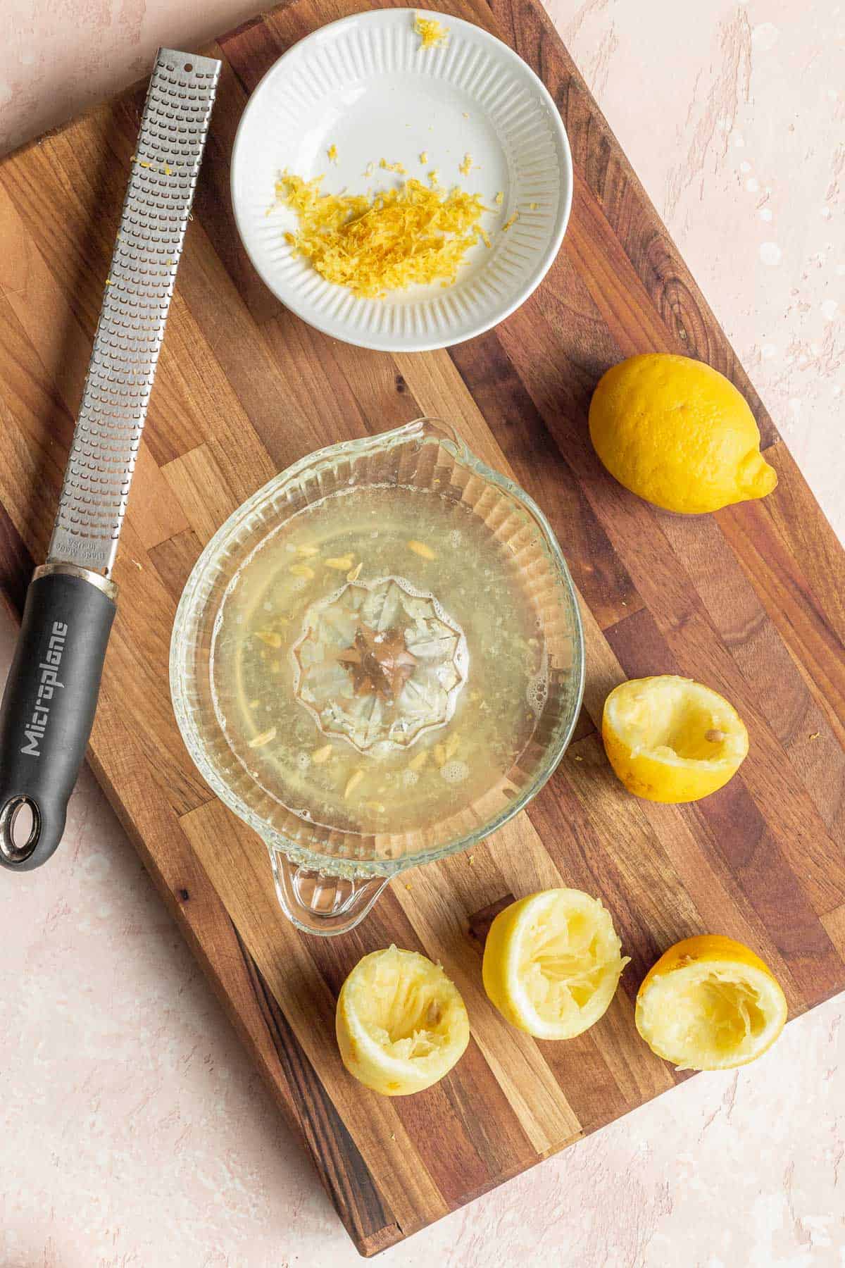 overhead shot of an old fashioned manual lemon juicer bowl filled with juice, a microplane, a plate of lemon zest, and several spent lemons on a wooden cutting board.