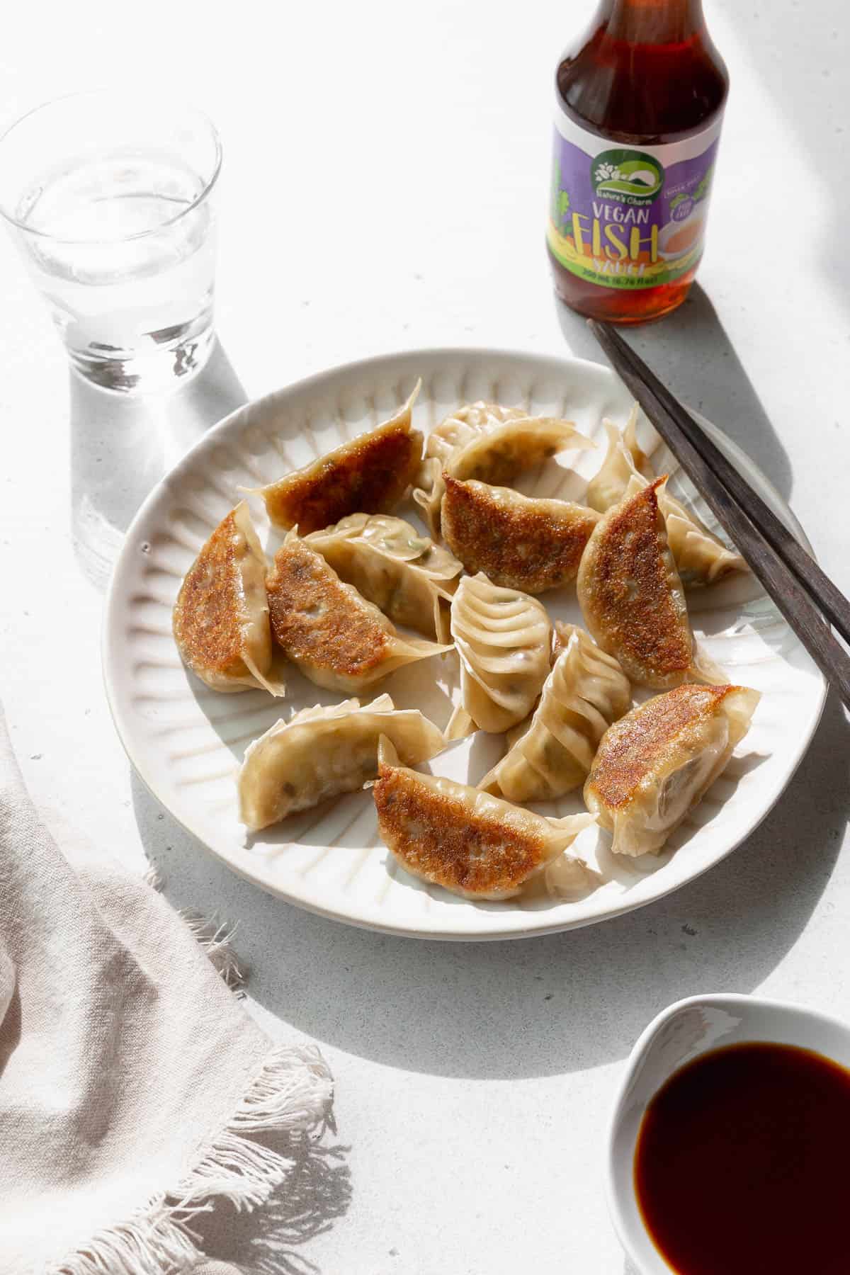 Angled shot of vegetable gyoza on a white plate with a glass of water and a bottle of vegan fish sauce in the background.