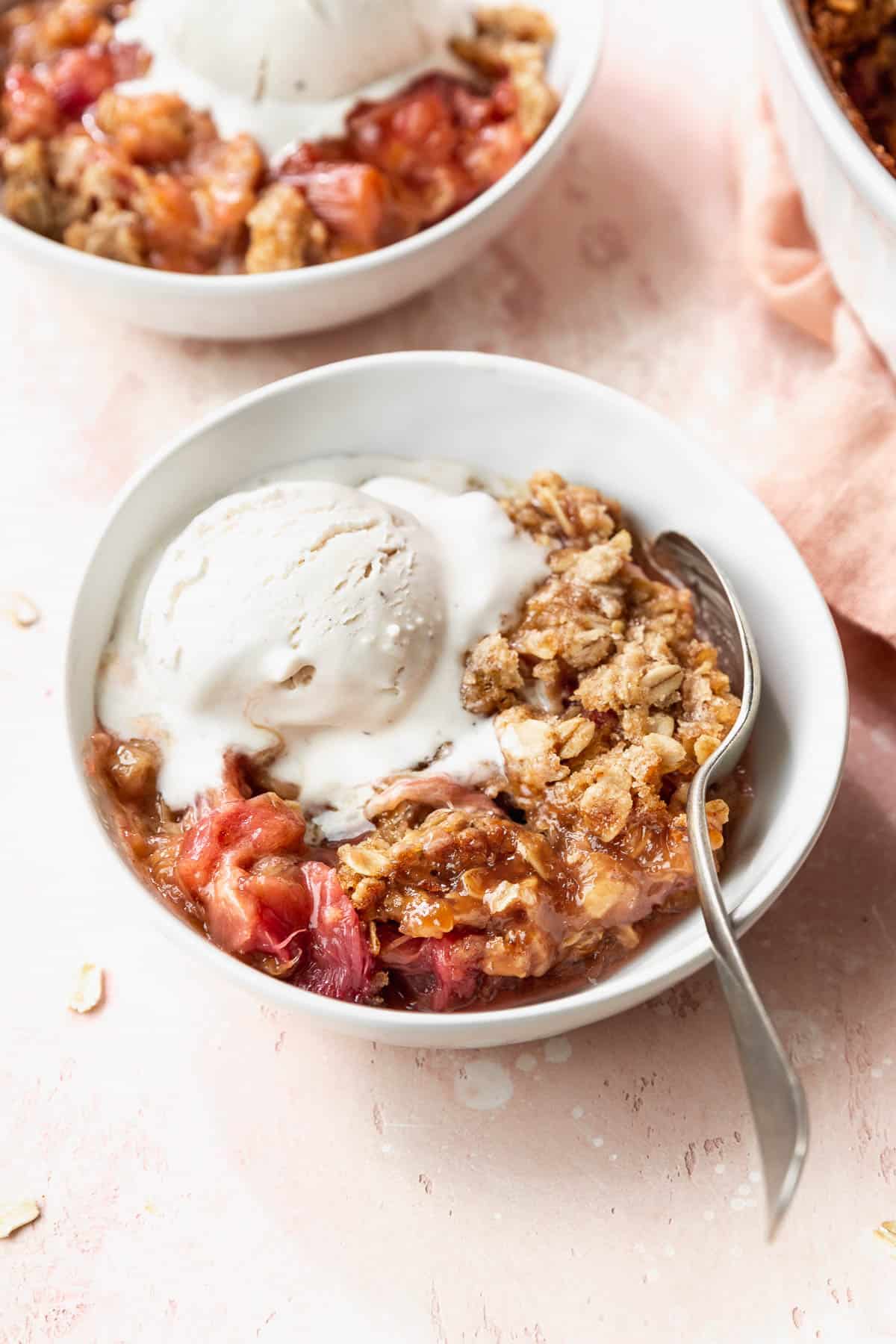 45 degree angle shot of a white bowl filled with gluten-free rhubarb crisp with oats crumble topping and a scoop of vanilla ice cream on a pink table.