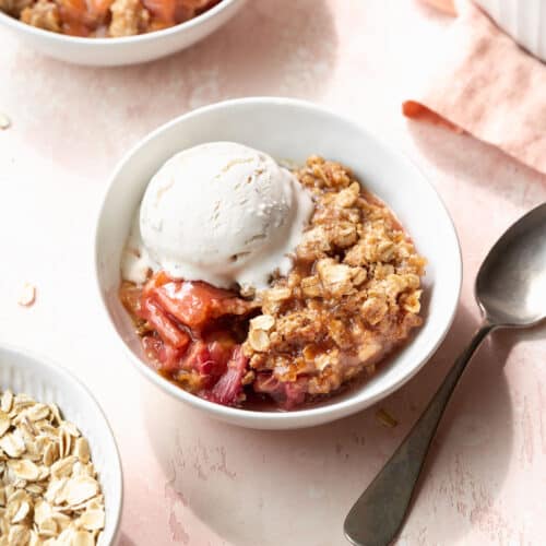 Square hero shot of a bowl of gluten-free rhubarb crisp in a white bowl with a scoop of vanilla ice cream and a silver spoon.