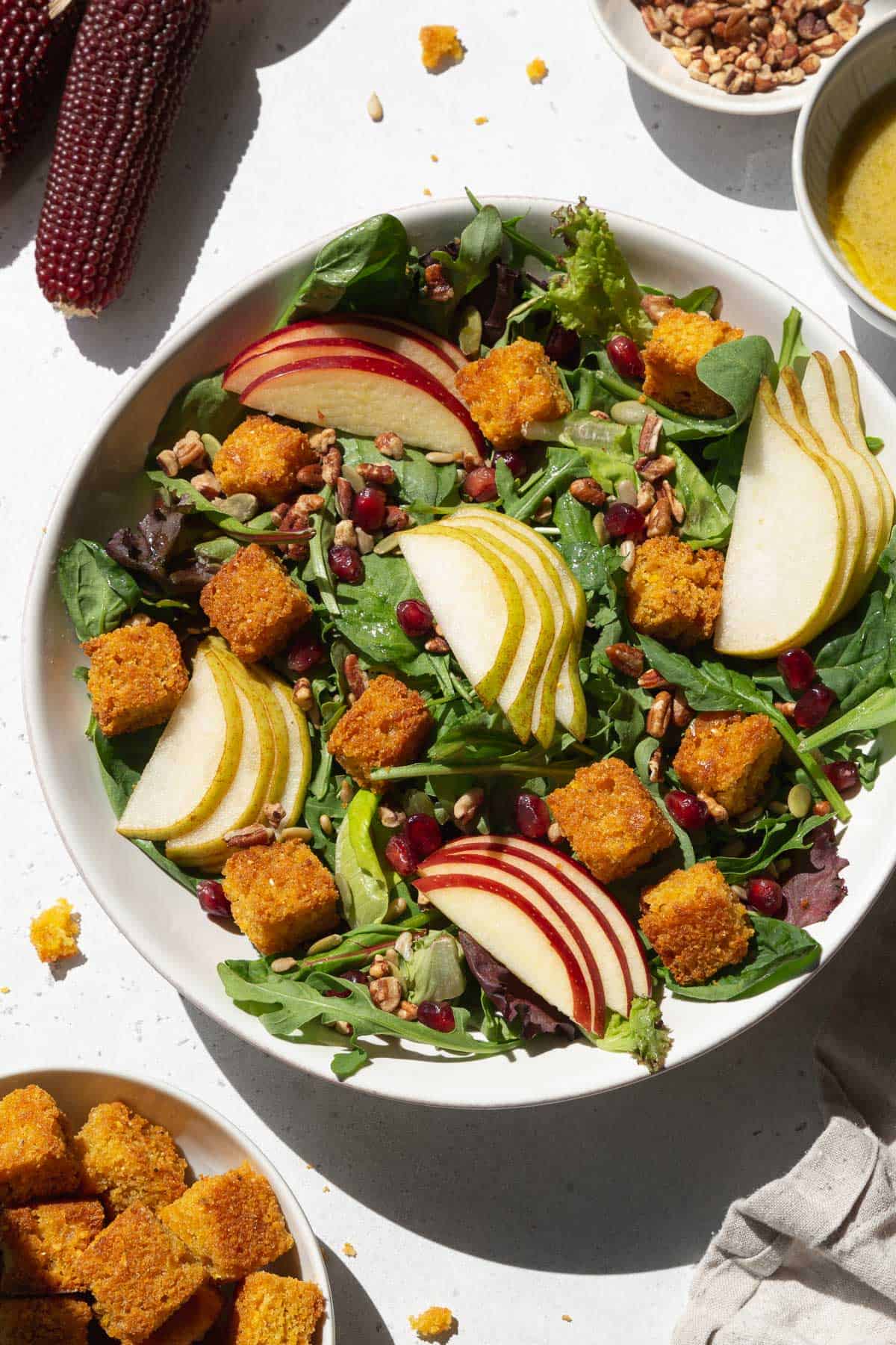 An overhead shot of a large white bowl of salad with cornbread croutons, sliced pears and apples, and pomegranate seeds.