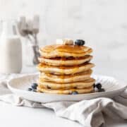 action shot of syrup being poured onto a stack of blueberry muffin mix pancakes topped with a pad of butter and extra fresh blueberries.