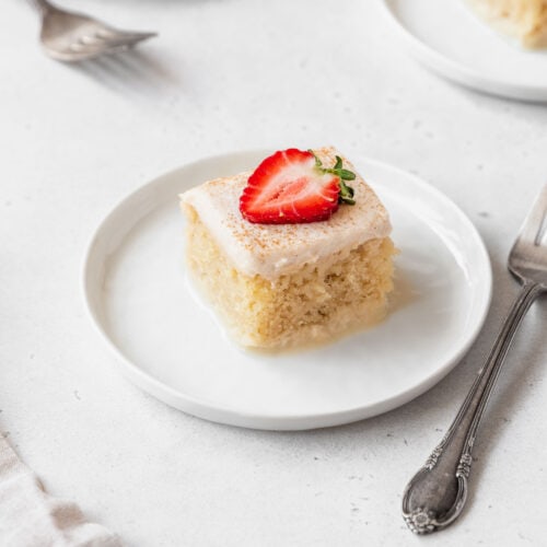 A square shot of a slice of gluten-free tres leches cake on a white dessert plate with a slice of strawberry on top.