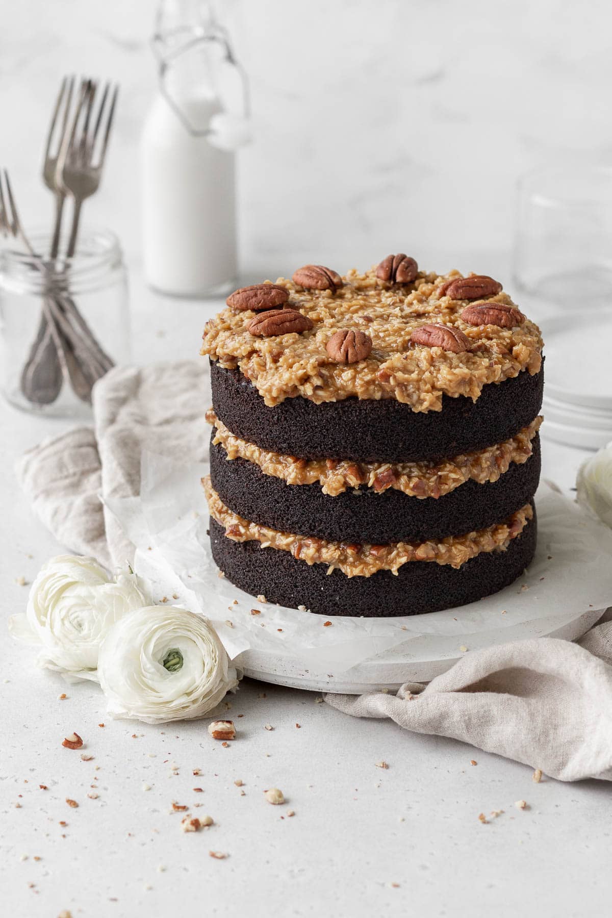 A three-layer German chocolate cake on a round white wooden board next to white flowers.