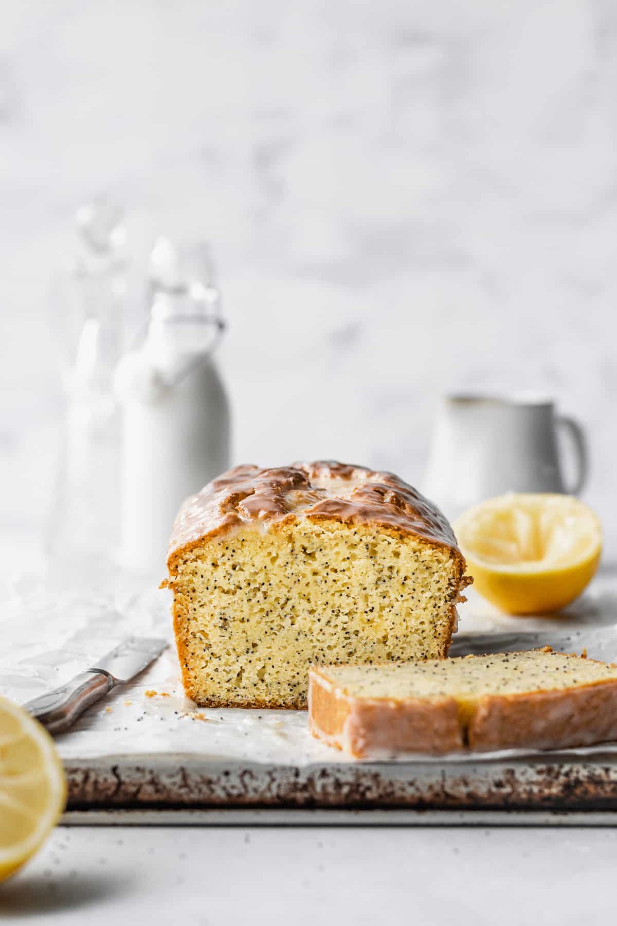 A sliced loaf of gluten free lemon poppy seed cake on a parchment paper with sliced lemons and milk jugs in the background.