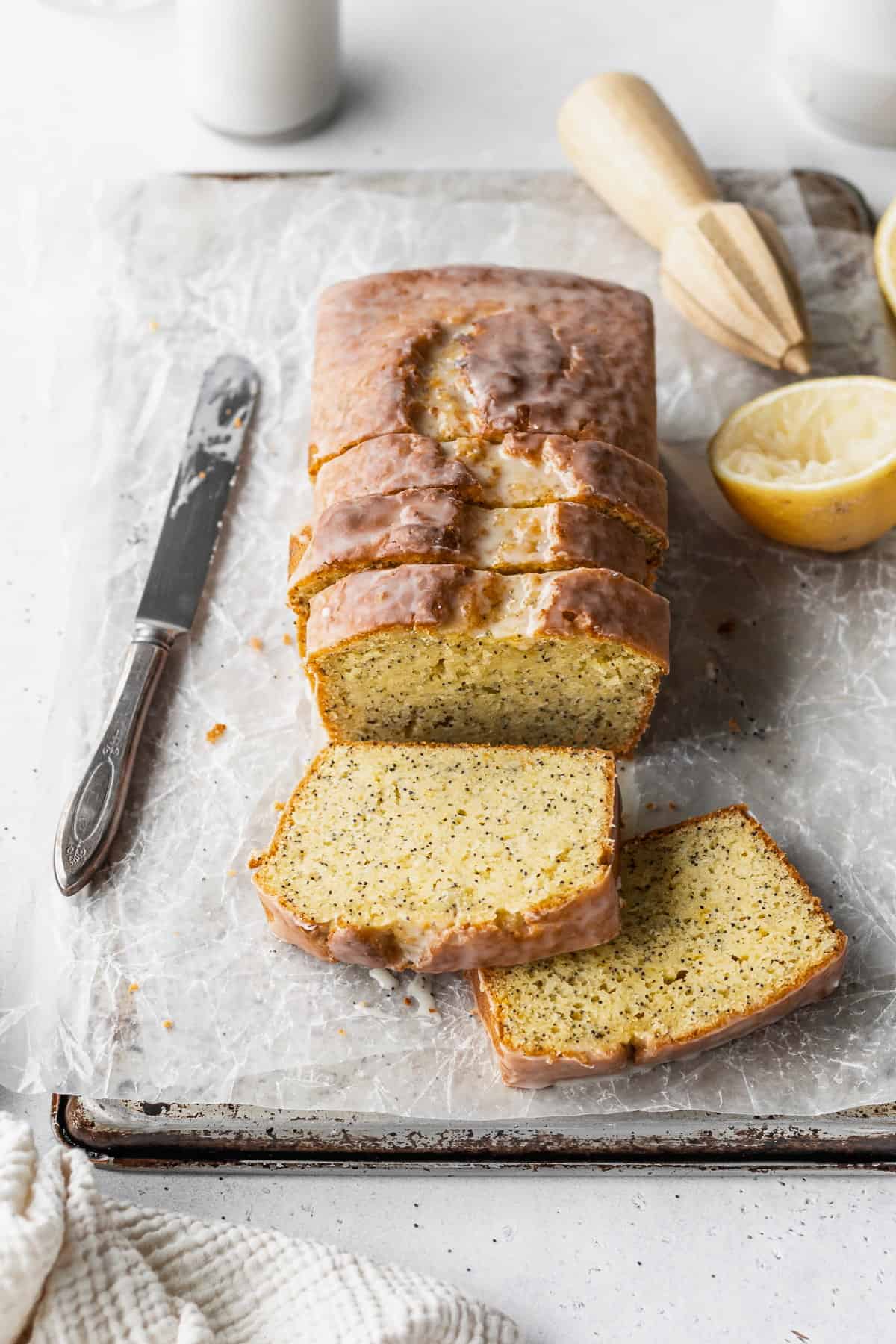 A sliced loaf of gluten-free lemon poppy seed loaf on a parchment paper lined baking sheet with a knife, half a lemon, and a wooden citrus zester placed around it.