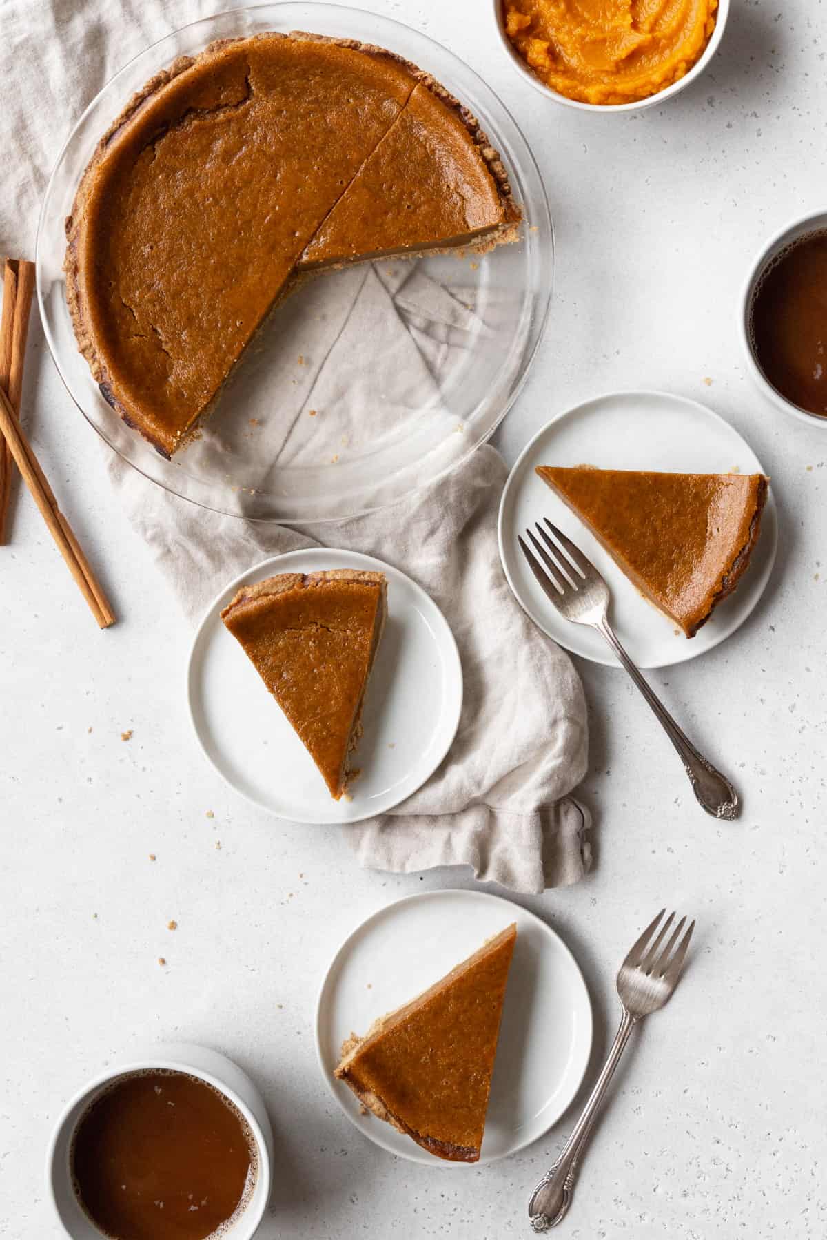 An egg free pumpkin pie in a glass pie plate with three slices on small white plates scattered on a white surface, with forks and mugs of coffee placed around them.