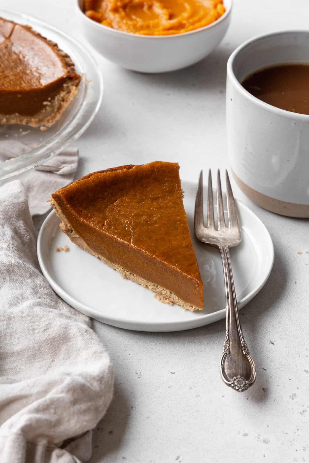 A slice of eggless pumpkin pie on a white dessert plate with a silver spoon, with a mug of coffee in the background.