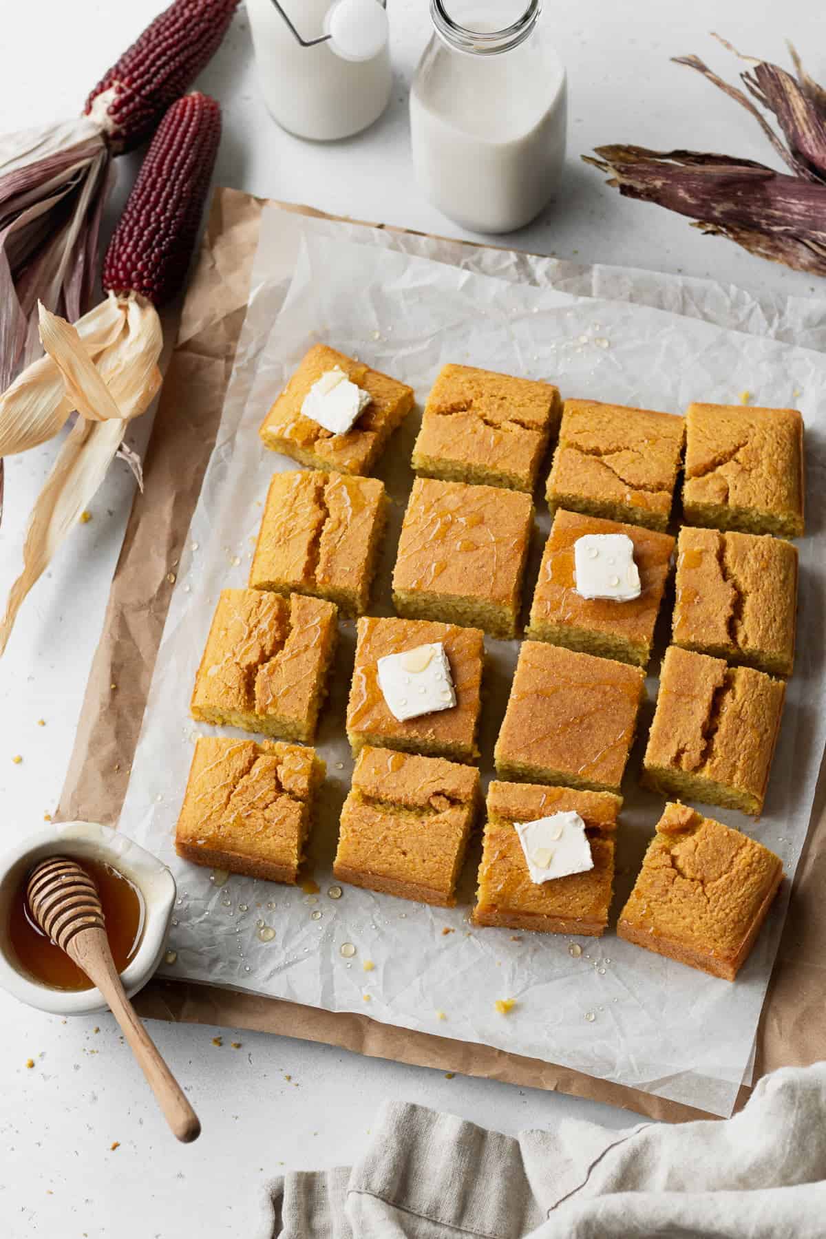 Table scene with dried corn on the cop surrounding a batch of dairy-free gluten-free cornbread cut into squares and topped with pads of butter.