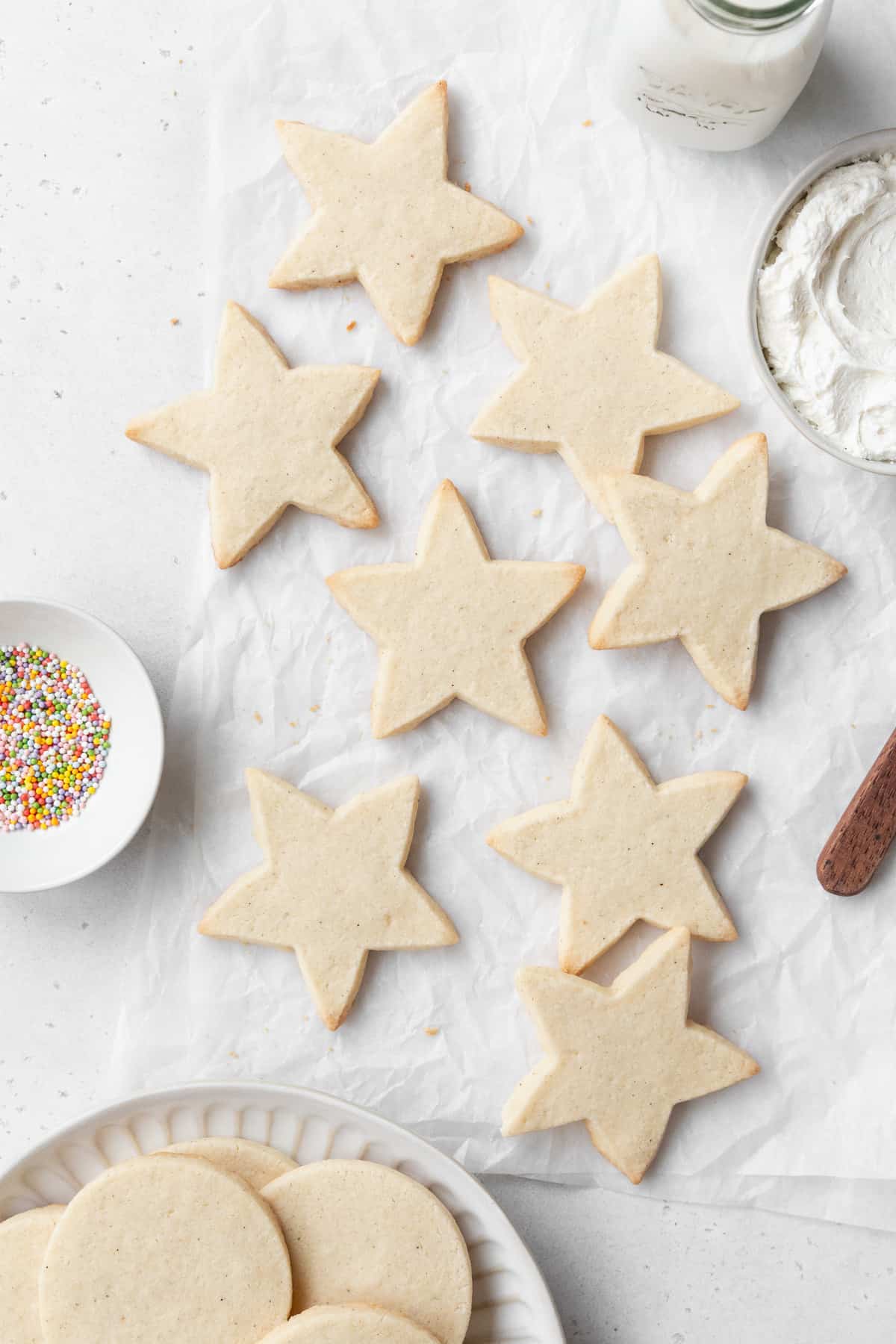 Star shaped gluten free sugar cookies on a white surface, with a small dish of sprinkles, a bowl of frosting, and a plate of circular sugar cookies on a plate surrounding them.