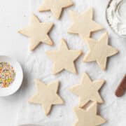 Star shaped gluten free sugar cookies on a white surface next to a small dish of sprinkles and a bowl of frosting.