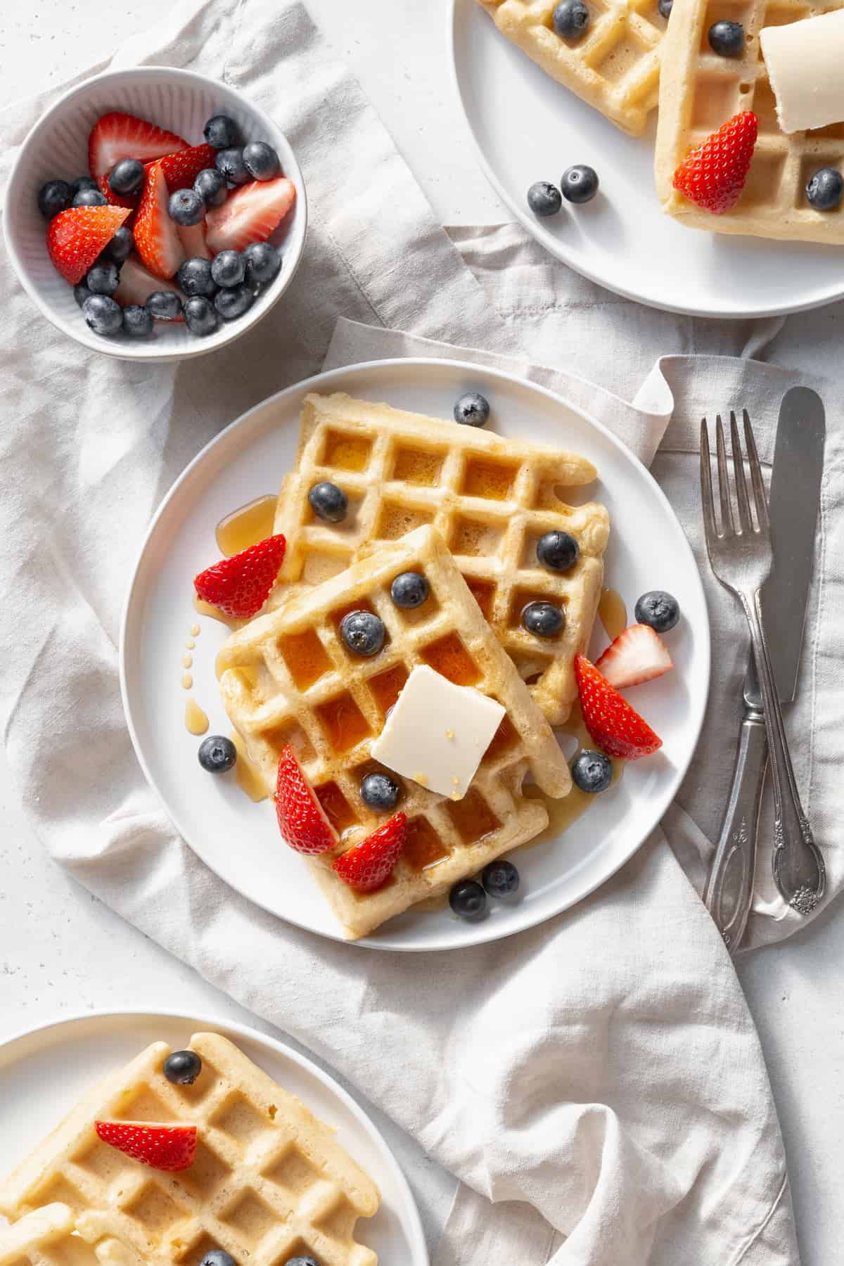 Plates of gluten-free waffles with butter and berries with a bowl of berries on the side.